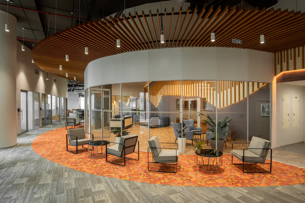 Office floor with a curved glass meeting room under timber ceiling slats and an orange-carpeted lounge