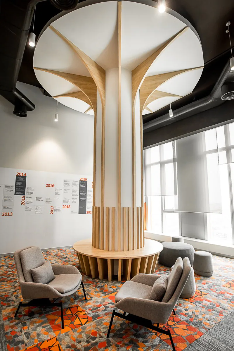 Office lounge built around a tree-like timber column with a round bench, grey armchairs and an orange patterned carpet