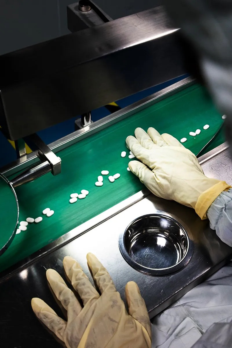 Gloved hands sorting white tablets on a green conveyor on a pharmaceutical packaging line