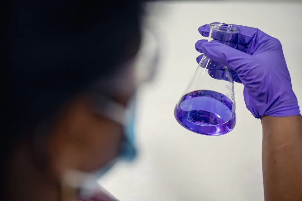 Gloved hand holding a conical flask of violet liquid, with a masked technician in the foreground