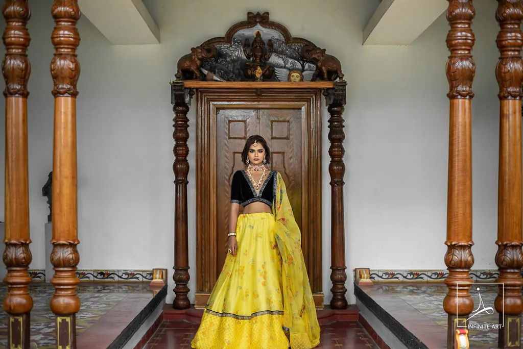 Model in a yellow lehenga and black velvet blouse standing before a carved wooden doorway between teak pillars