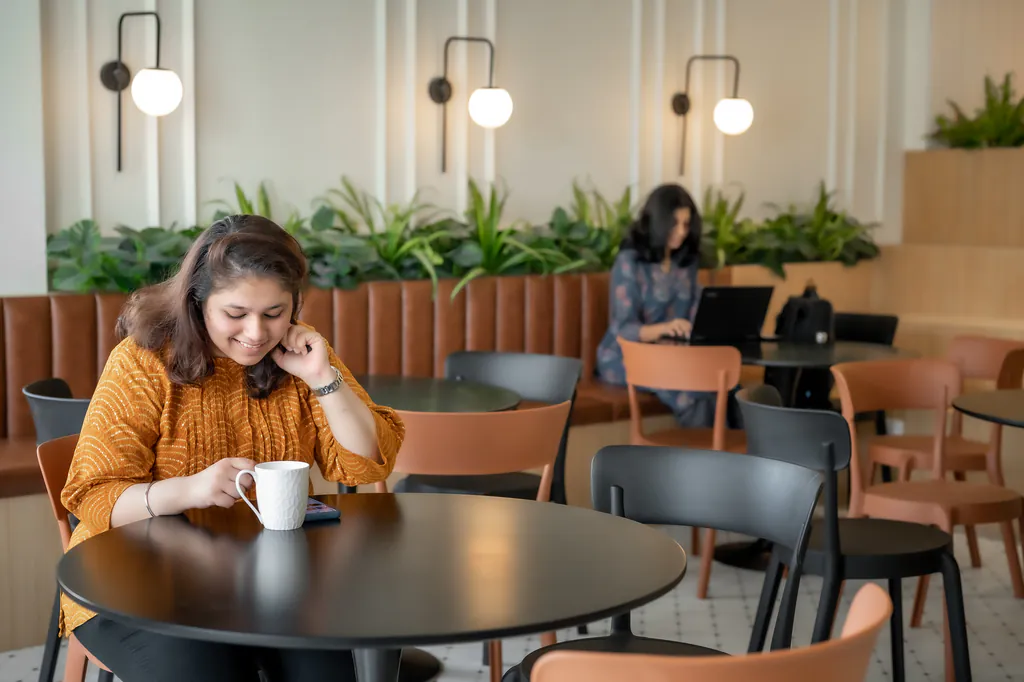 Woman with a coffee cup at a round table in a co-working café, with another person working on a laptop behind her