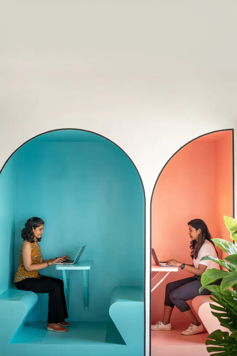 Two people working on laptops inside arched turquoise and coral booths in an office