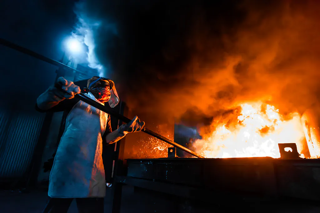 Worker in a protective suit and visor stirring a furnace as flames and sparks rise