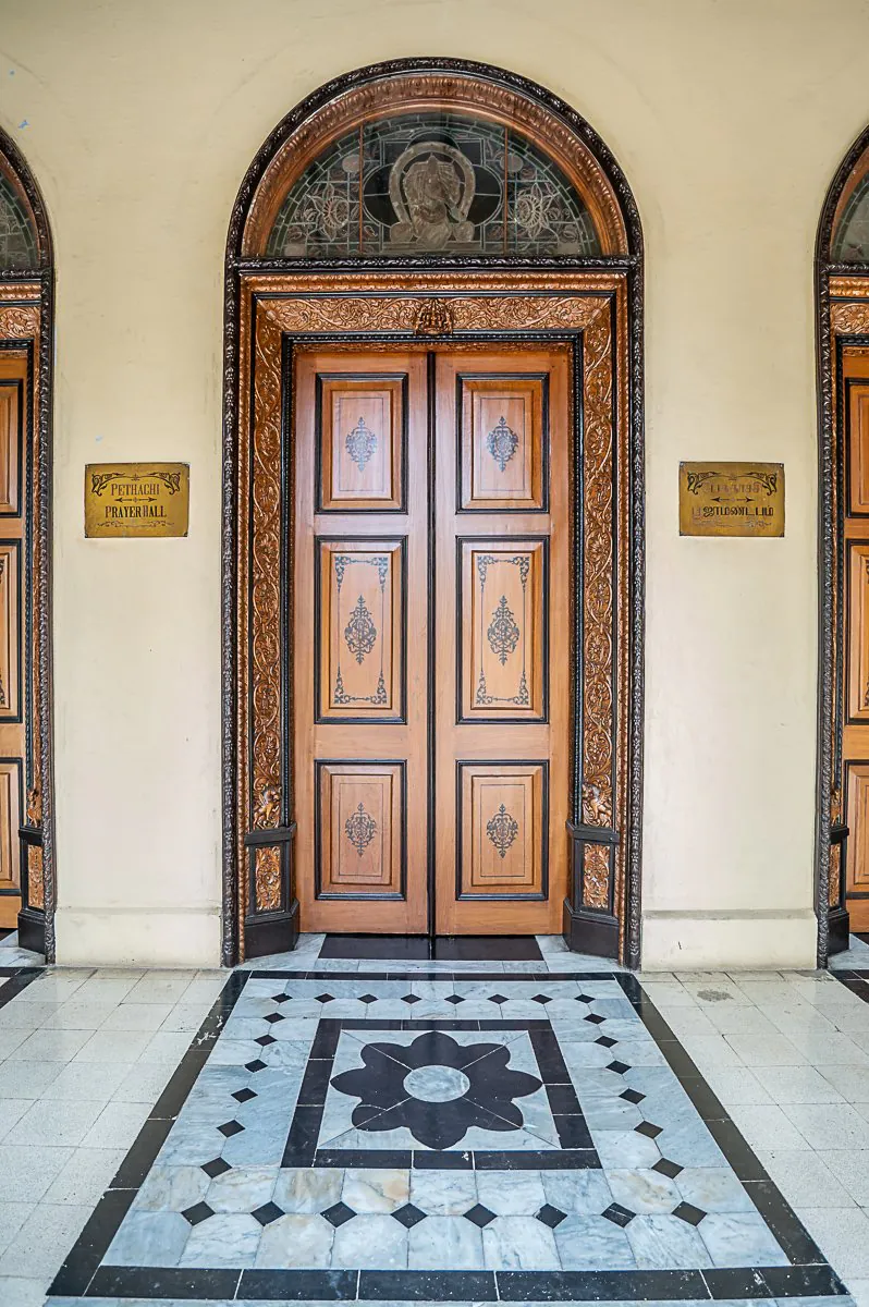 Carved teak doors under a stained-glass arch at the Pethachi Prayer Hall, with a black-and-white marble floor