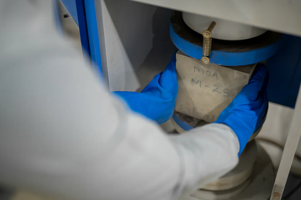 Gloved hands placing a concrete test cube into a compression testing machine