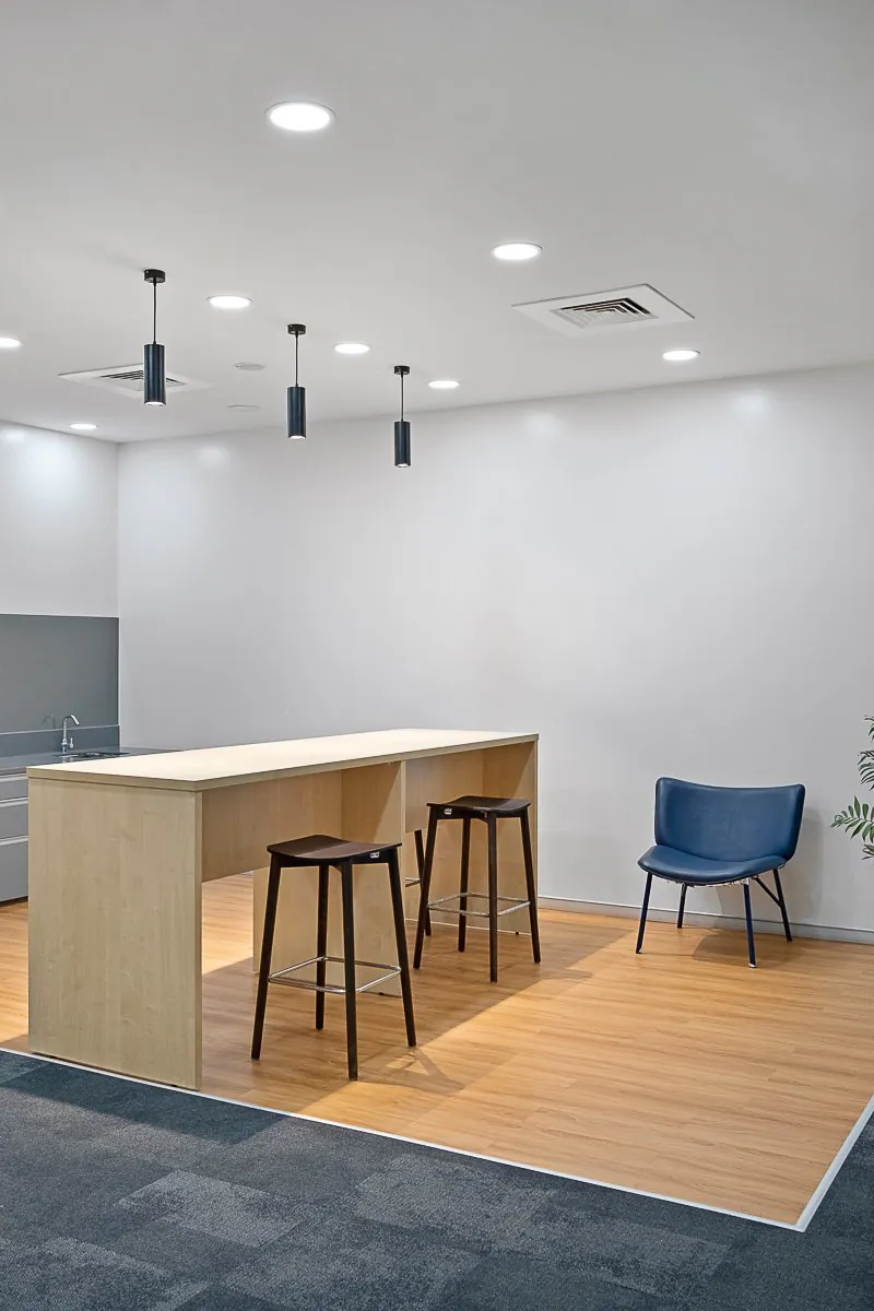 Office pantry with a light-wood high table, two bar stools, a blue lounge chair and black pendant lights