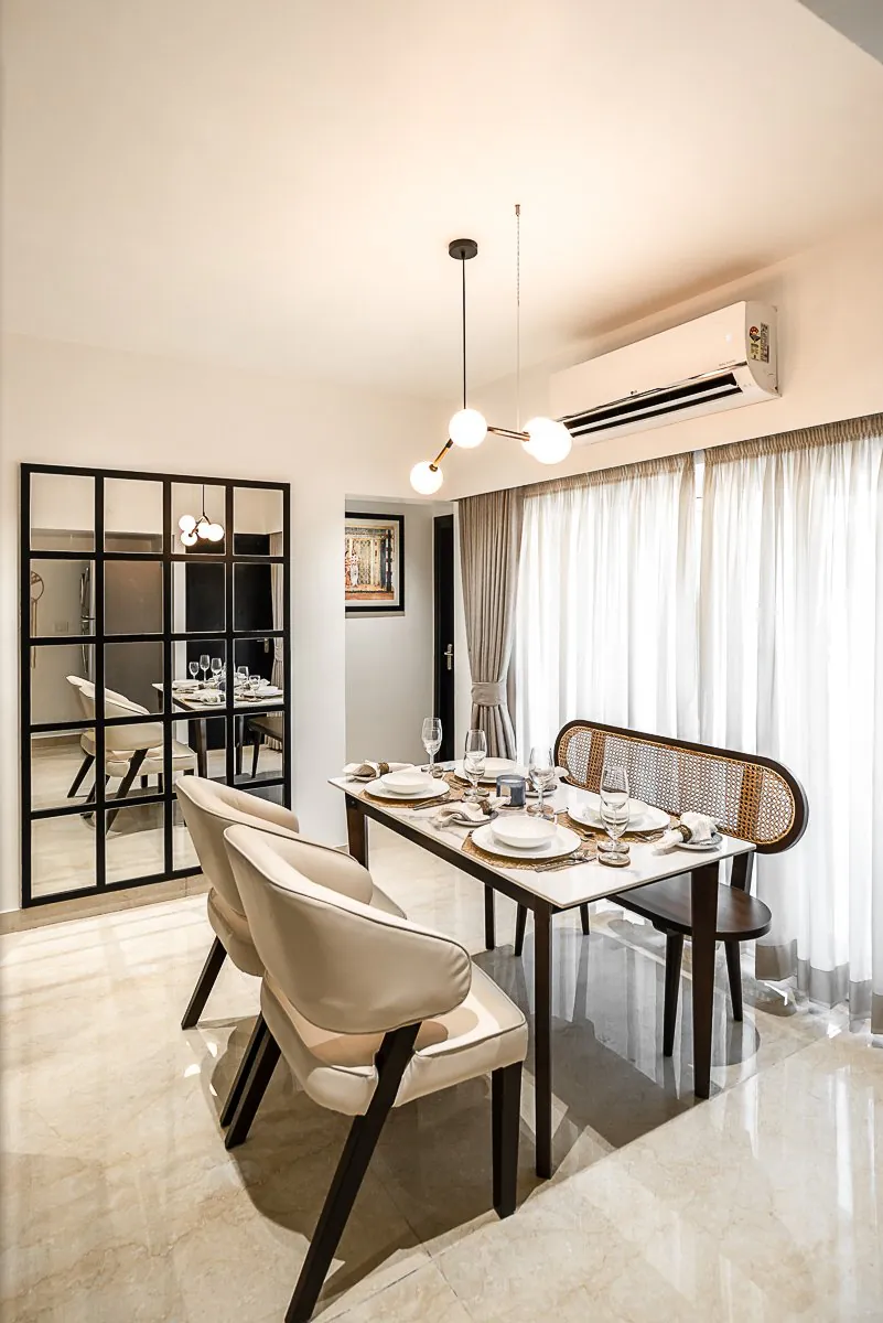 Dining table with a cane-back bench and cream chairs under a globe pendant, beside a black grid mirror