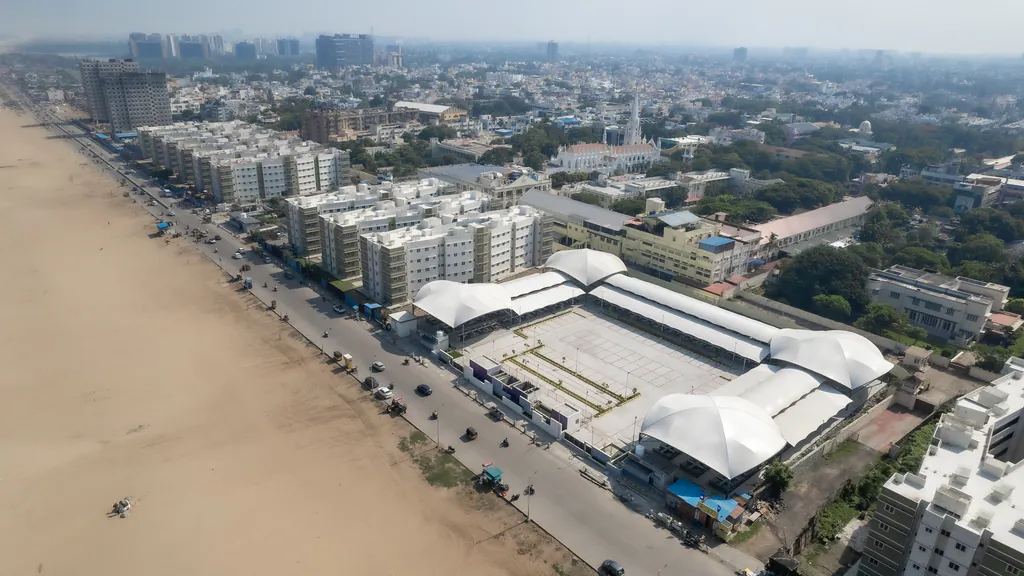 Drone view of a white tensile-roof structure beside a beach and coastal road, with the city behind