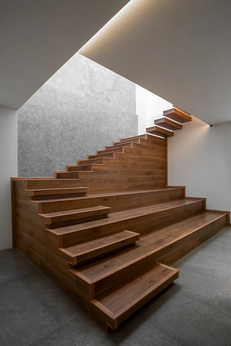 Timber staircase with floating treads rising beside a textured grey wall under a skylight